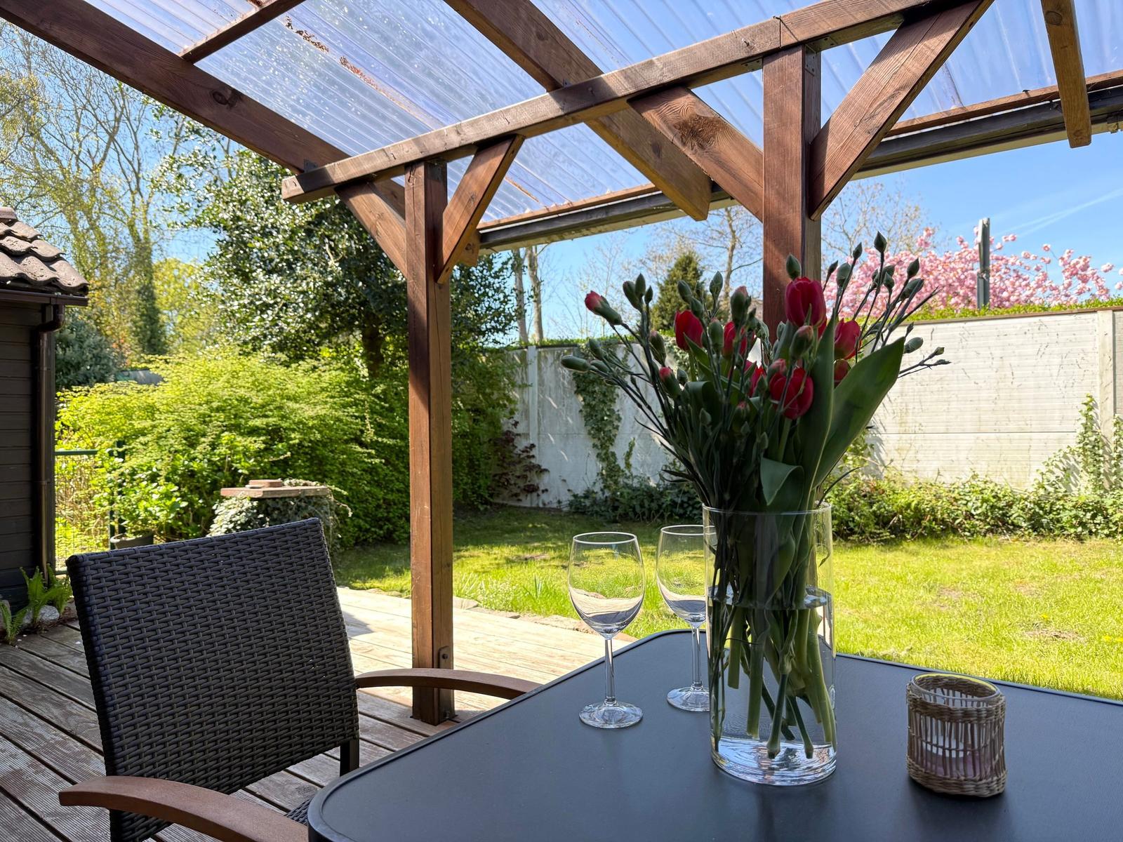 Tisch mit Blumen und Gläsern unter einer Holzveranda mit Blick auf den Garten.