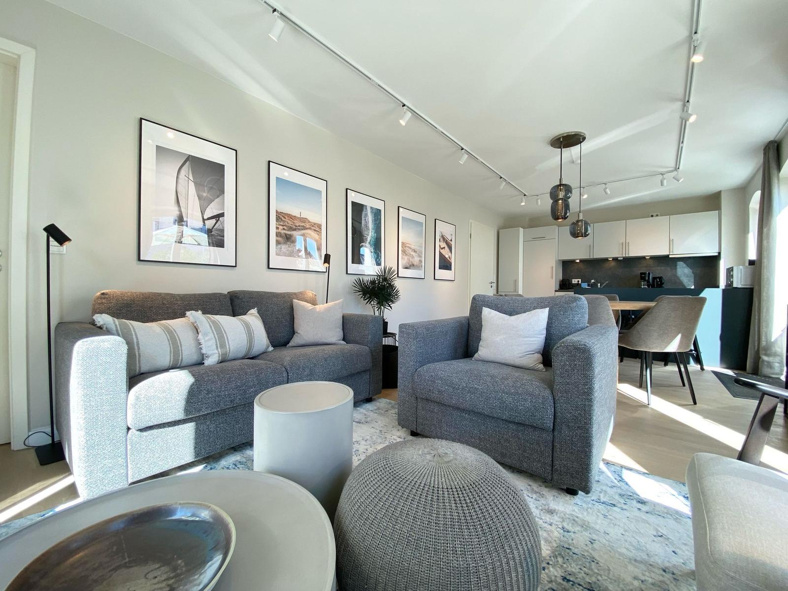 Wohnbereich mit grauem Sofa, Sessel und Wandbildern. Küche im Hintergrund.