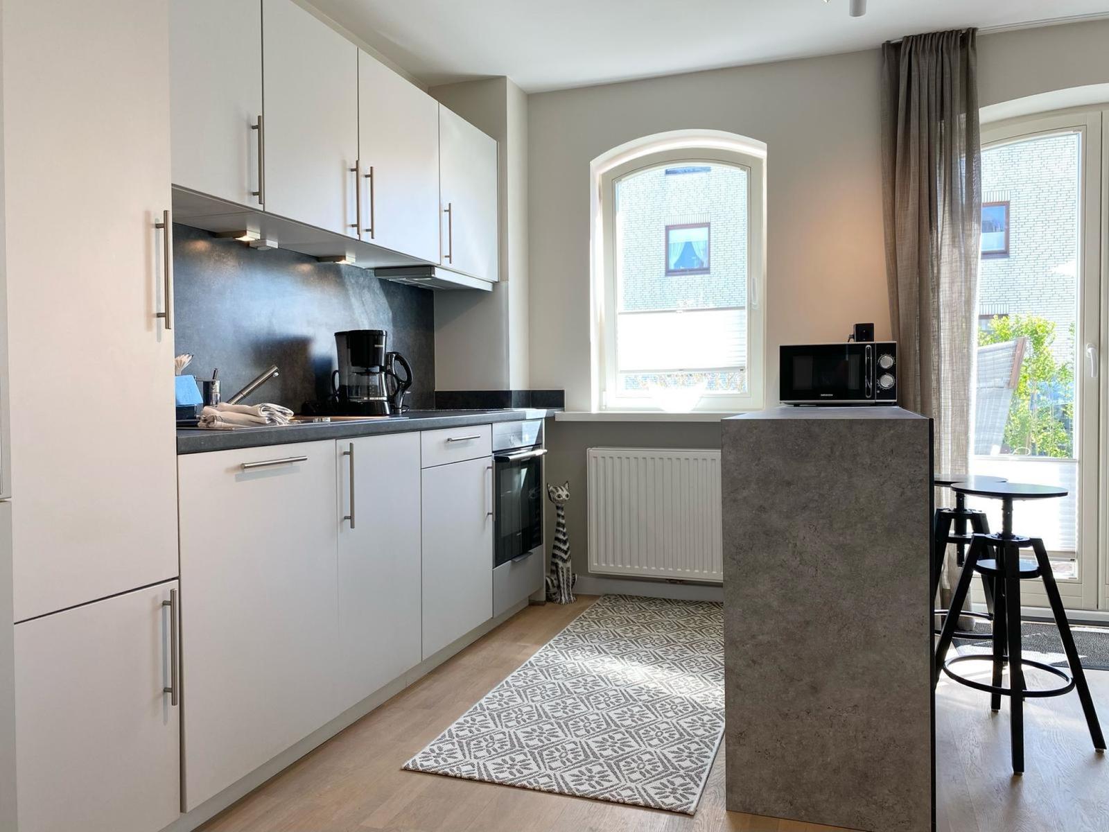 Küche mit weißen Schränken, Arbeitsplatte und Fenster. Es steht ein Mikrowellenherd und Barhocker.