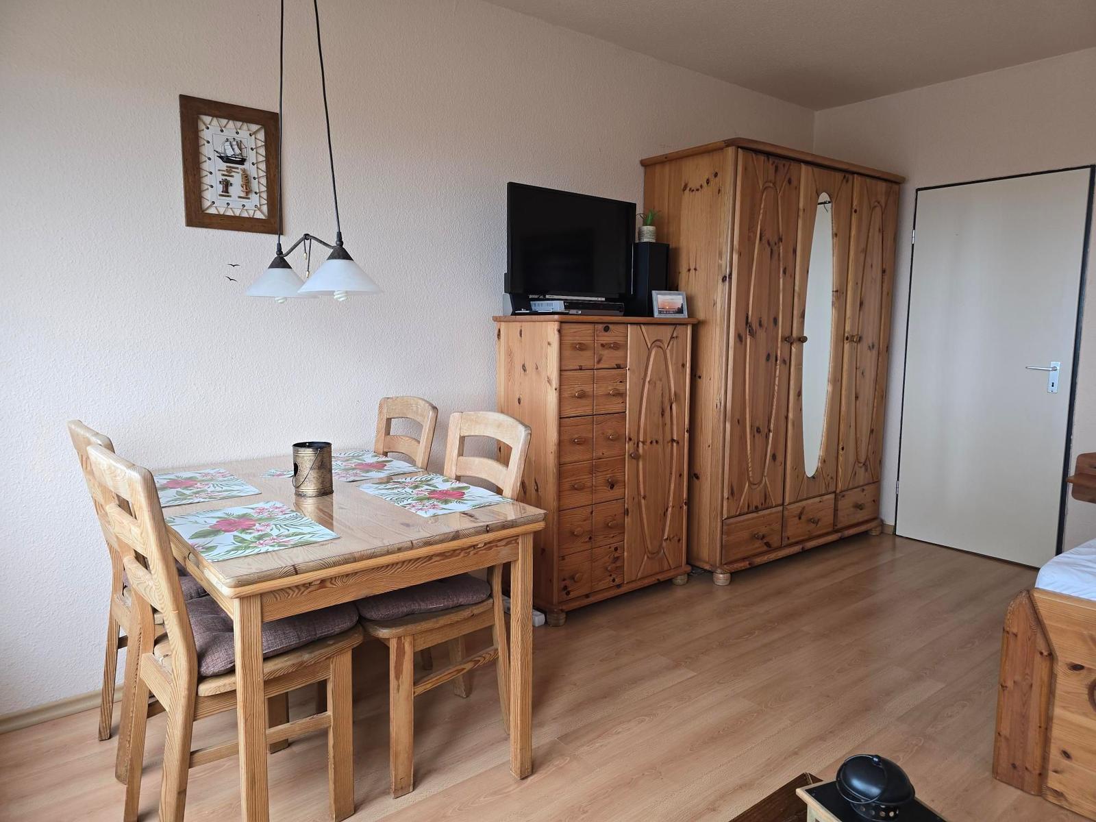 Essbereich mit Holztisch, Stühlen und TV. Holzschrank und Tür im Hintergrund.