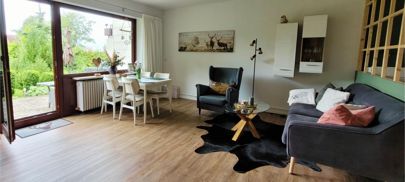 Wohn- und Essbereich mit Sofa, Sessel und Tisch. Durchgang zur Terrasse mit Gartenblick.