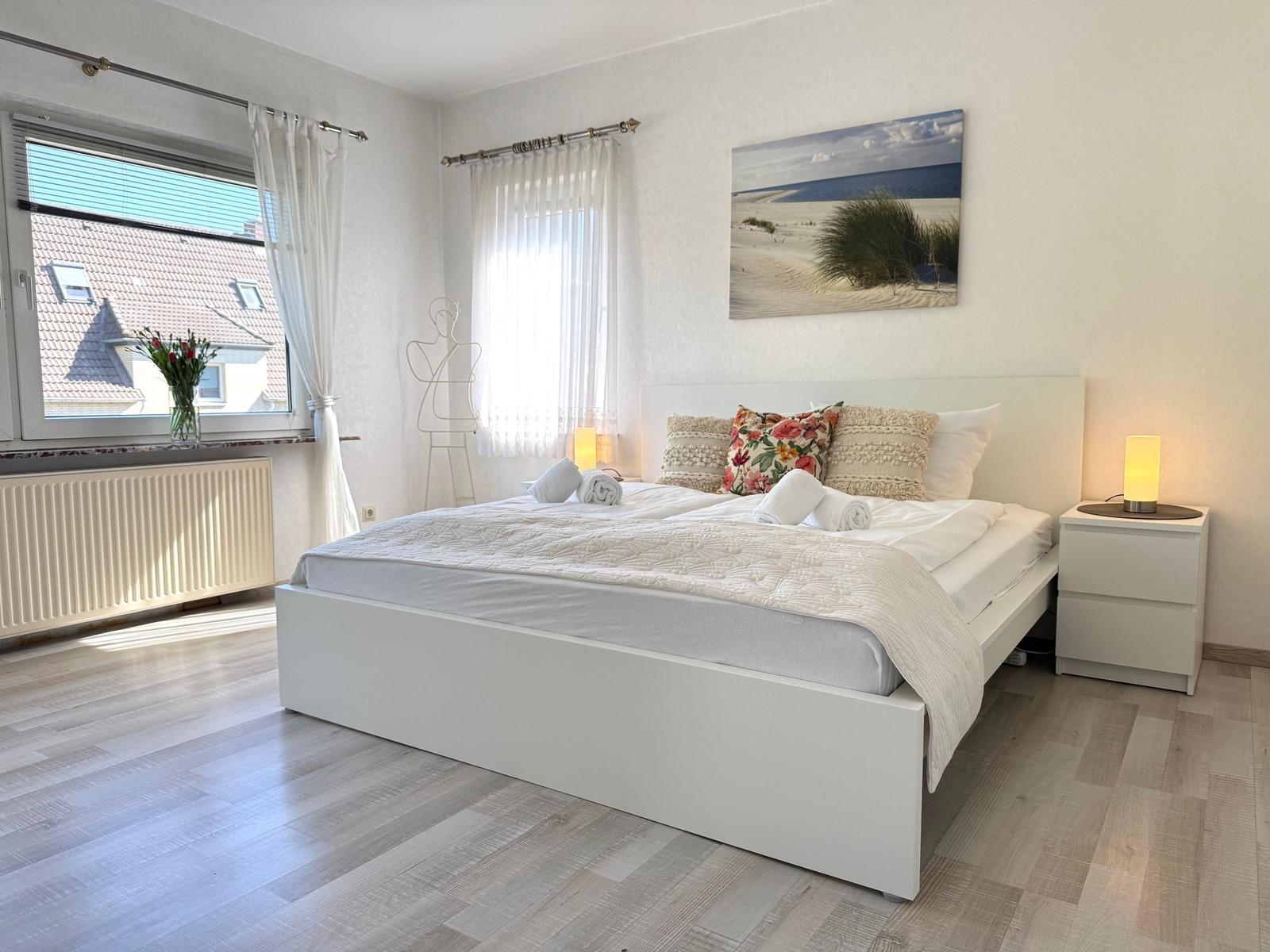 Schlafzimmer mit weißem Bett, Nachttisch und Fenster mit Blick auf Dächer.