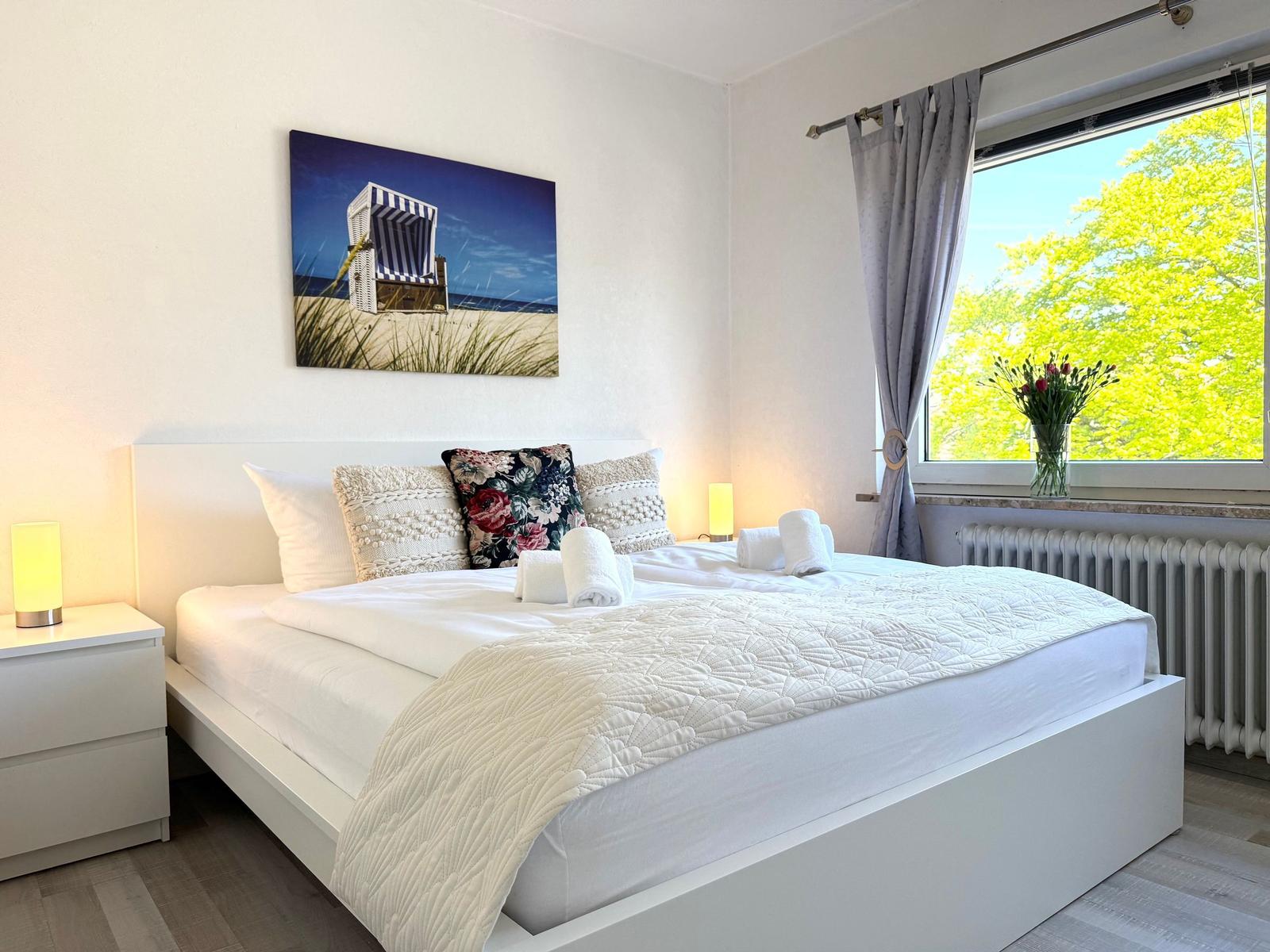 Schlafzimmer mit weißem Bett, Wandbild und Blick auf Bäume.