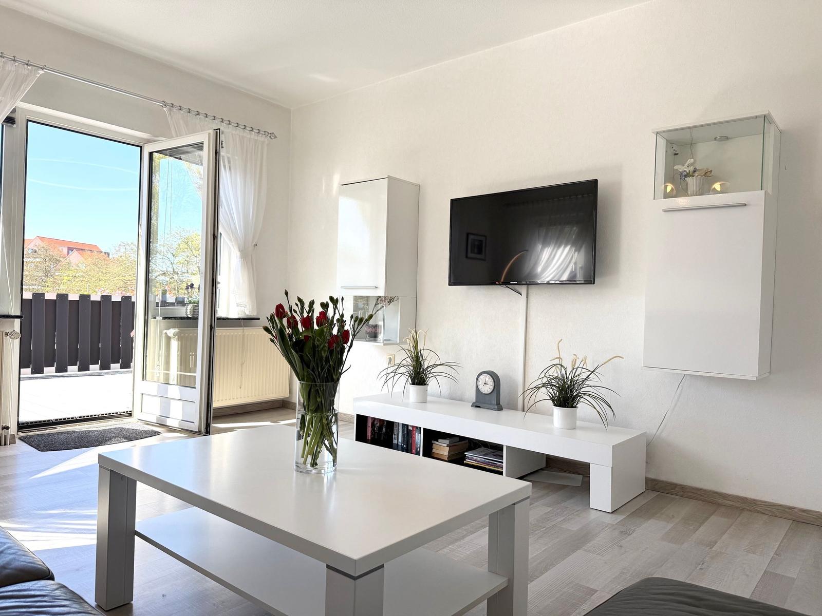 Wohnzimmer mit Fernseher, Tisch und Balkontür. Weiße Möbel und Pflanzen.