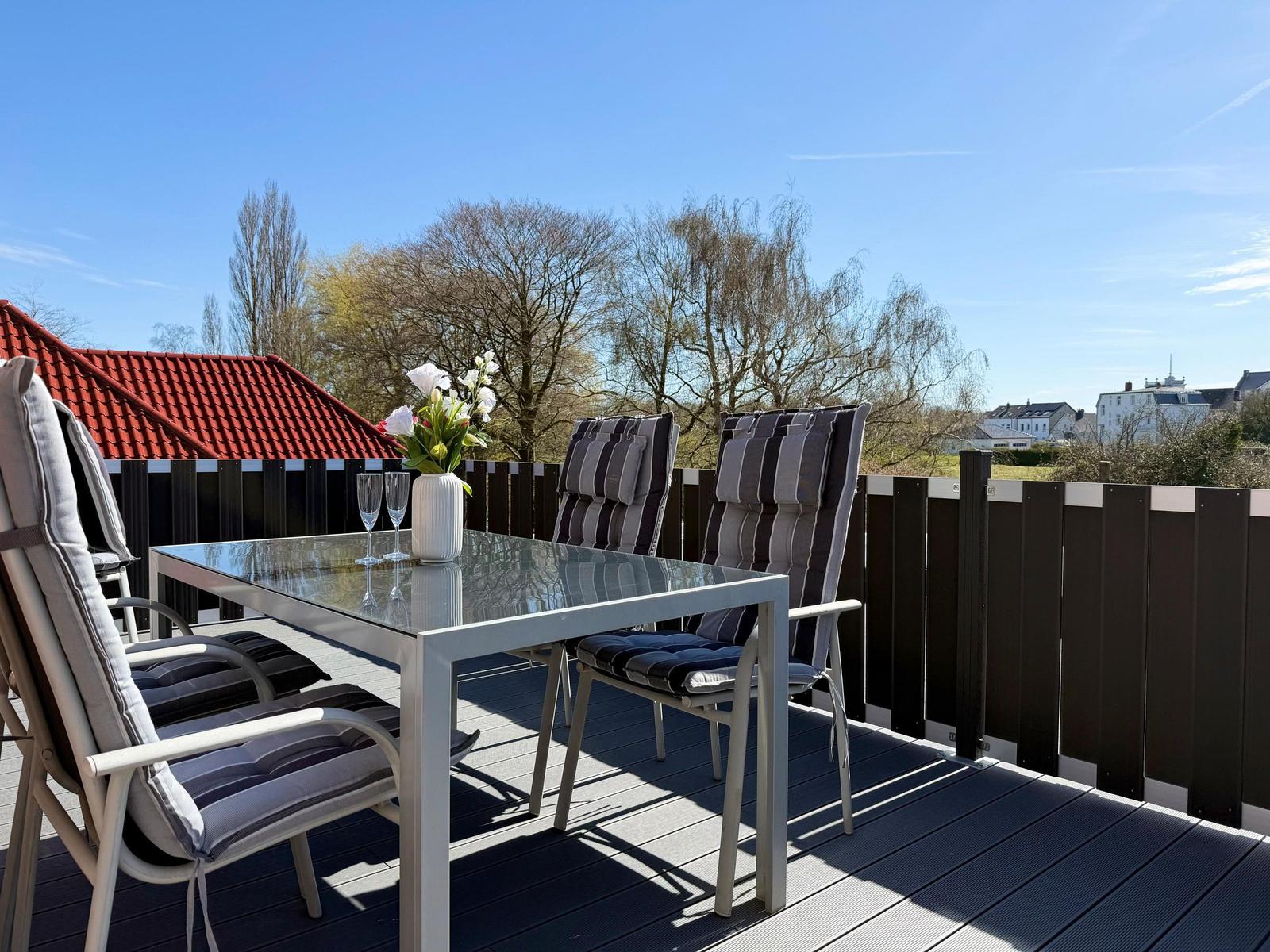 Terrasse mit Glas-Tisch, Stühlen und Blumen. Blick auf Bäume und Gebäude.