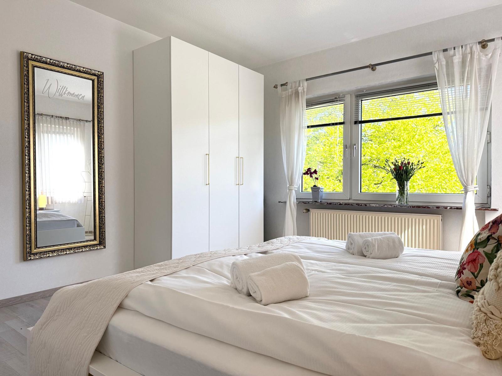 Schlafzimmer mit weißem Bett, Schrank und Spiegel. Fenster mit Blick auf grüne Bäume.