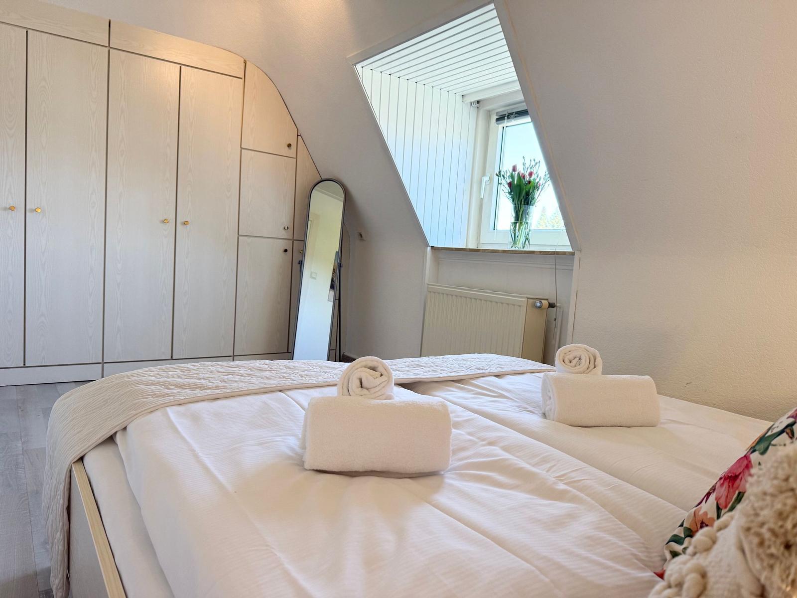 Schlafzimmer mit Dachschrägen, Bett, Kleiderschrank und Fenster mit Blumen.