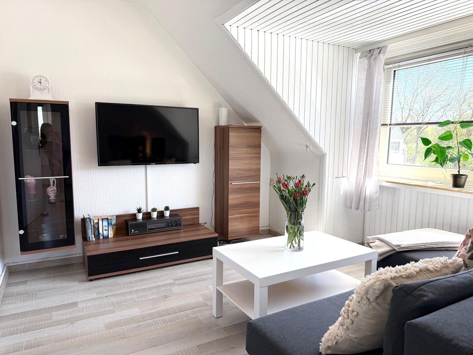 Wohnzimmer mit TV, Couch, Tisch und Fenster. Dachgeschoss mit Holzboden und weißen Wänden.