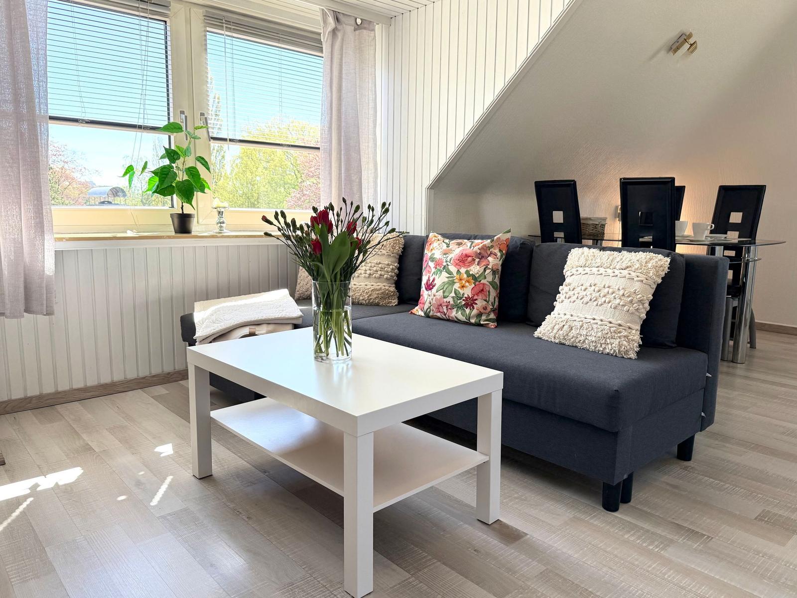 Ein Wohnzimmer mit Sofa, Tisch und Fenster. Im Hintergrund ist ein Esstisch mit Stühlen.