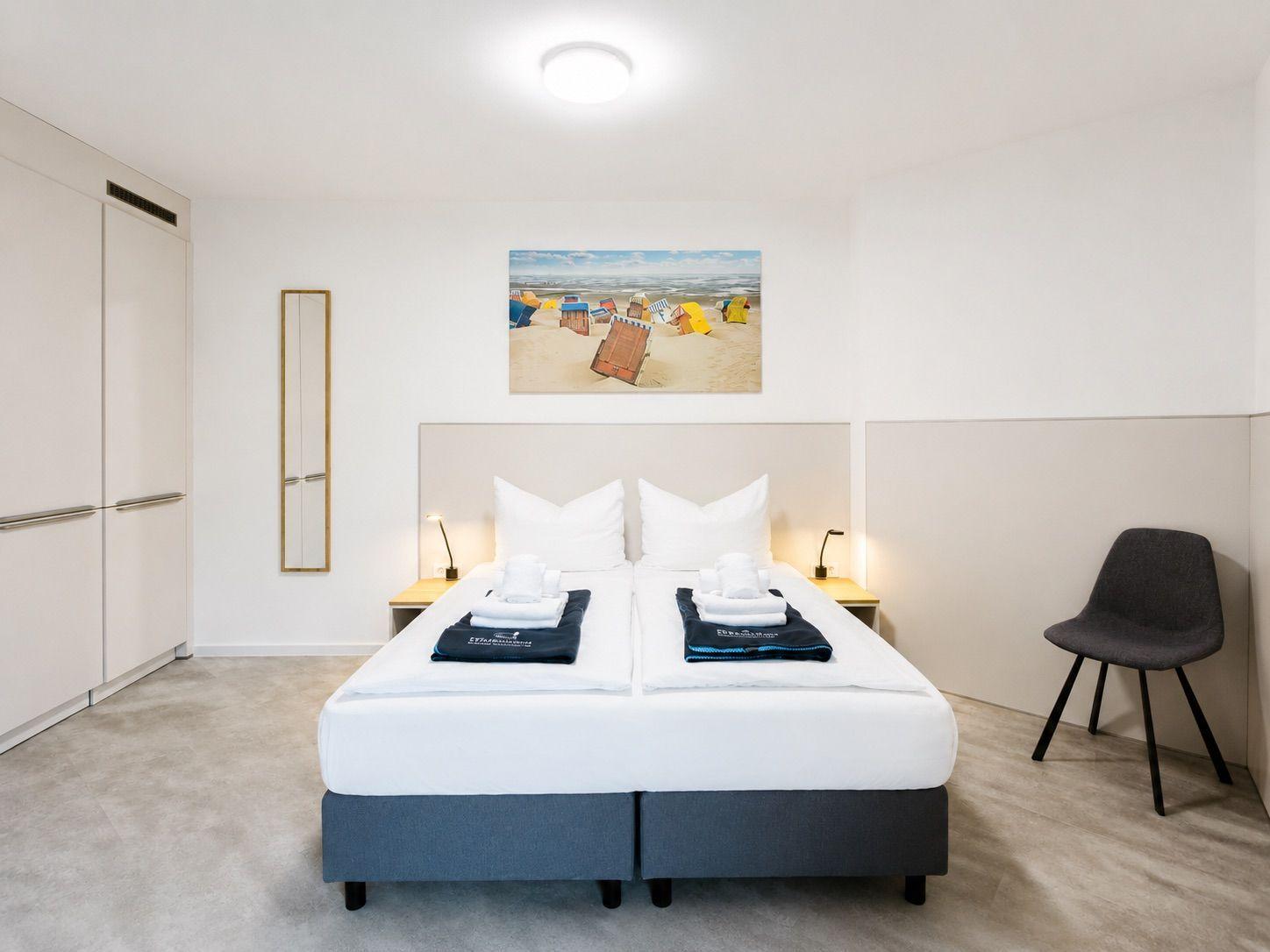 Schlafzimmer mit Doppelbett, Kleiderschrank und Stuhl. Wandbild zeigt Strand.