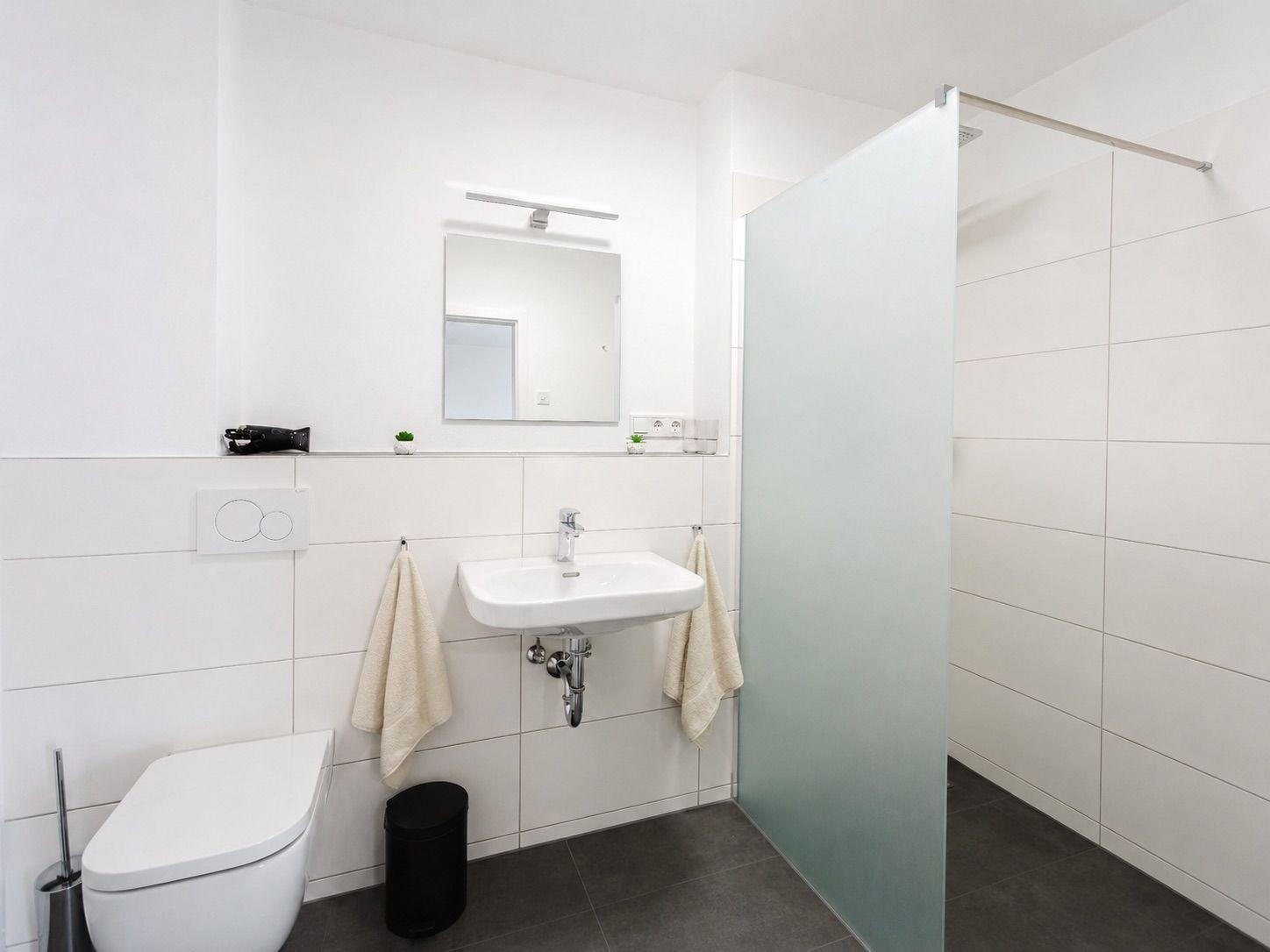 Bad mit Dusche, Waschbecken und Toilette. Weiße Fliesen und dunkle Bodenfliesen.