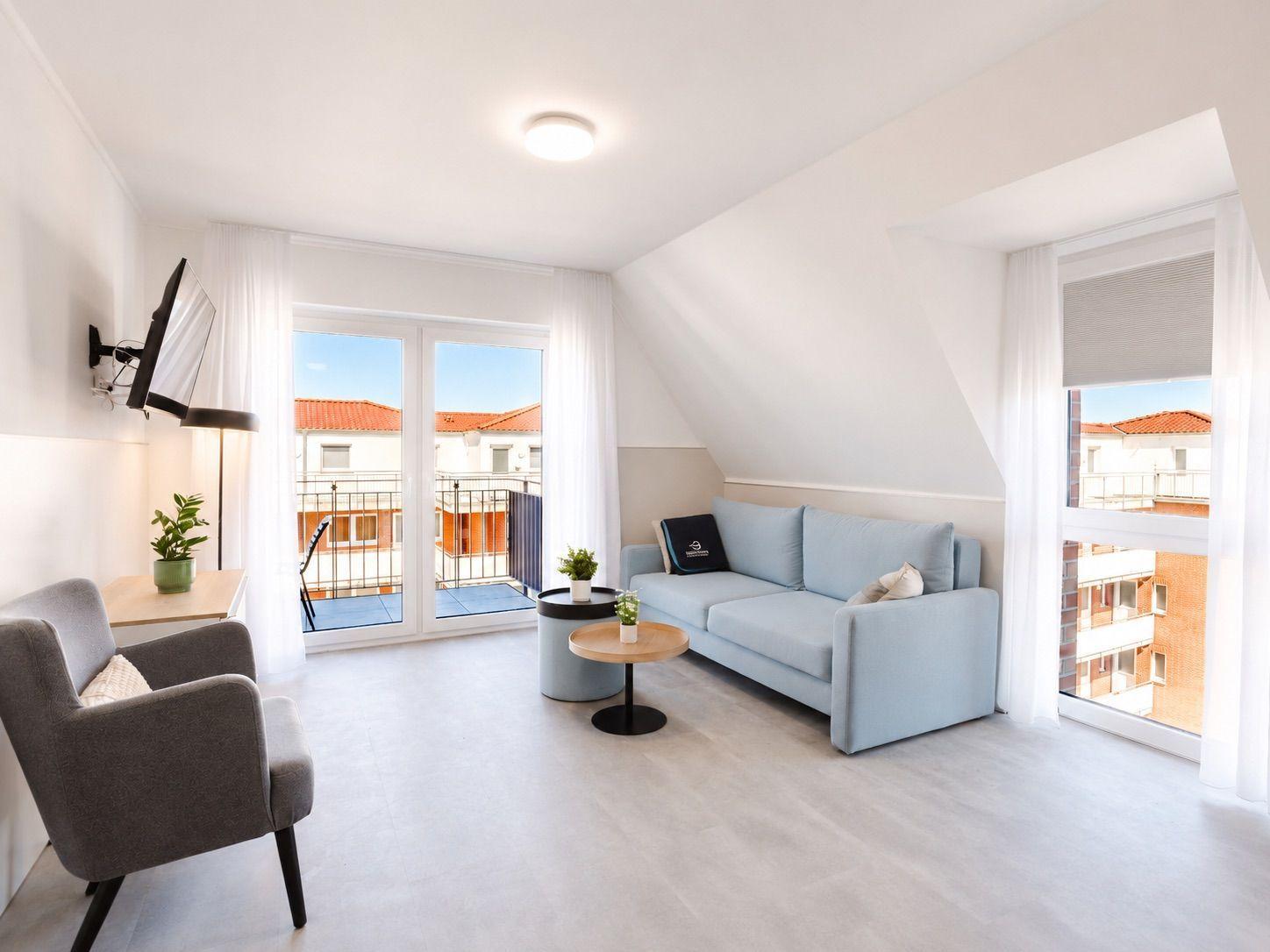 Living room with sofa, armchair, and balcony. Windows overlook buildings.