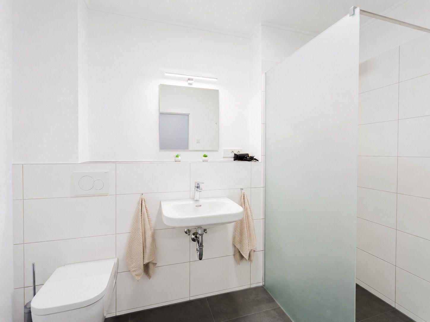 Badezimmer mit Dusche, Waschbecken und Toilette. Weiße Fliesen und dunkle Bodenfliesen.