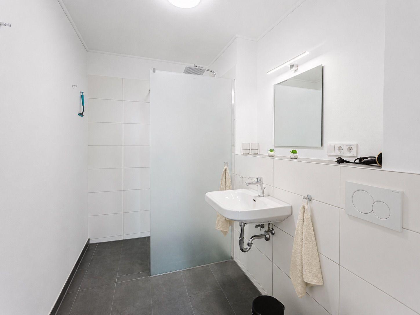 Badezimmer mit Dusche, Waschbecken und Spiegel. Weiße Fliesen und graue Bodenfliesen.