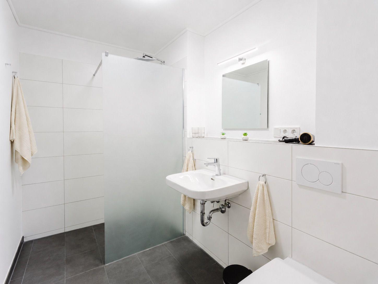 Badezimmer mit Dusche, Waschbecken und Toilette. Weiße Wände, grauer Boden und Spiegel.
