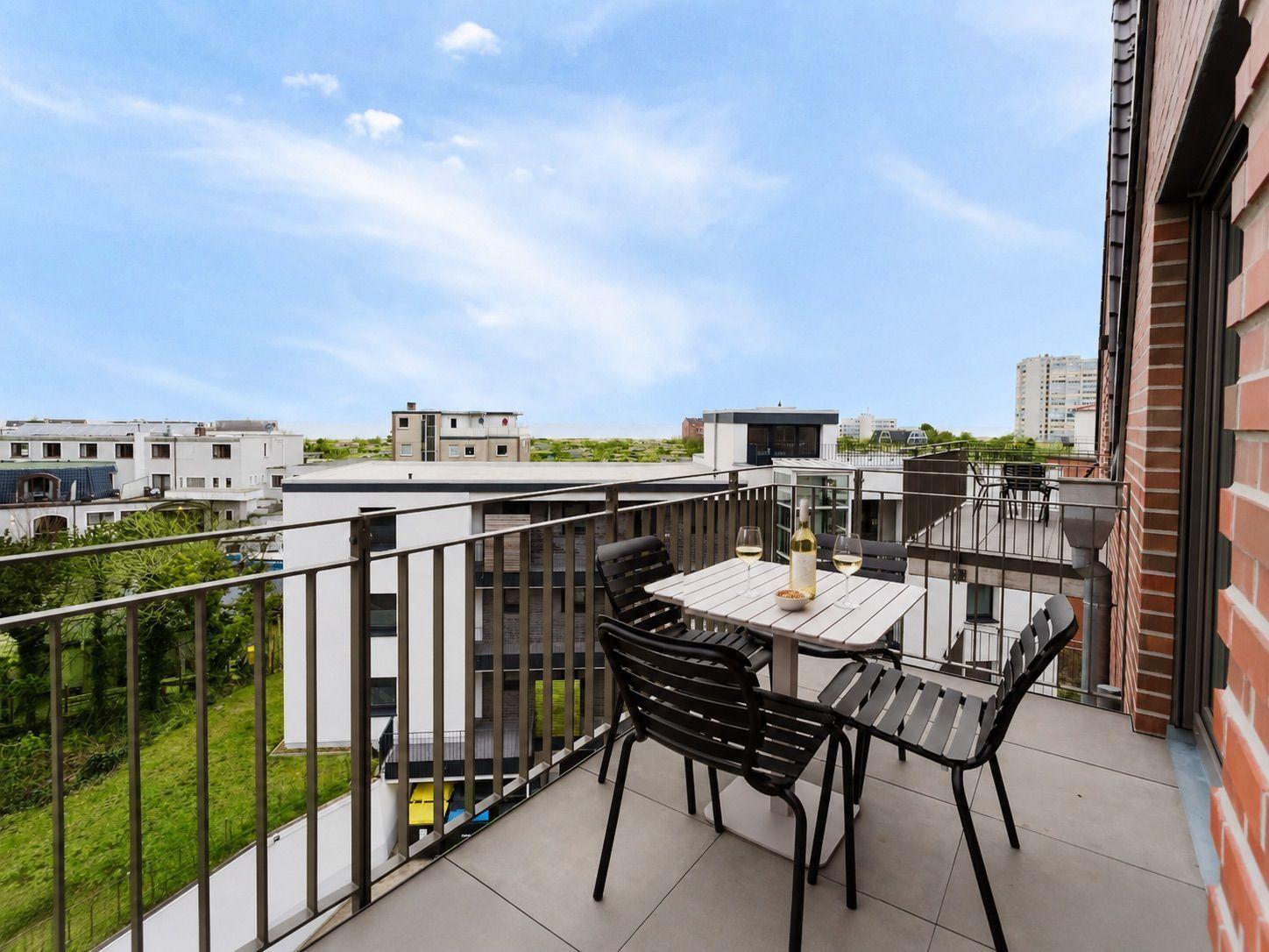 Terrasse mit Tisch und Stühlen, Blick auf Gebäude und Grünflächen unter blauem Himmel.