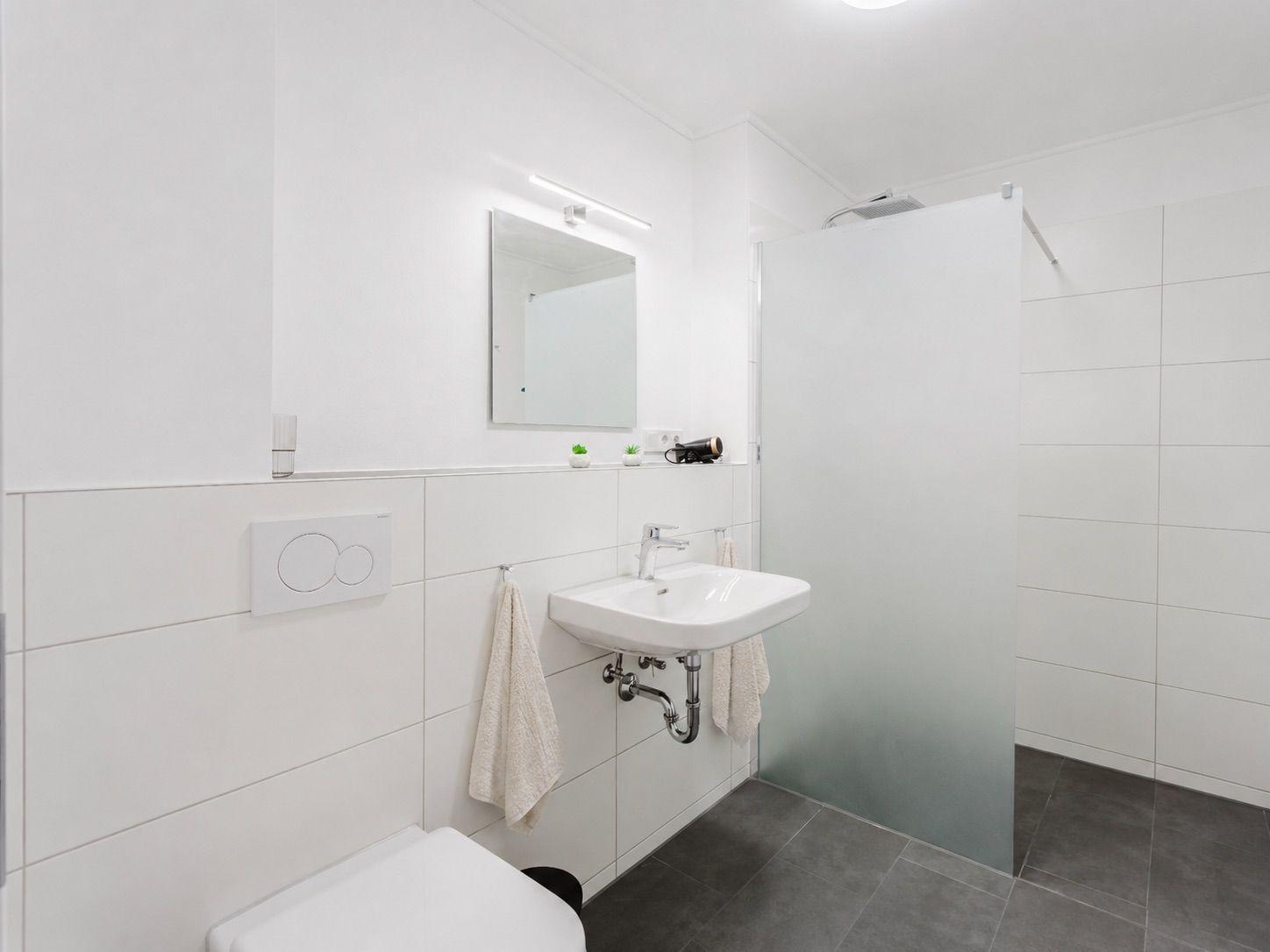 Badezimmer mit Dusche, Waschbecken und Toilette. Weiße Fliesen und graue Bodenfliesen.