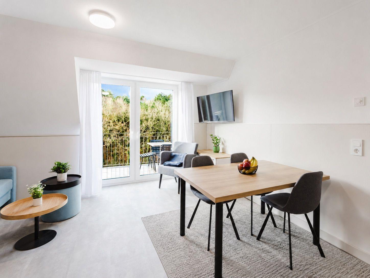 Wohn- und Essbereich mit Tisch, Stühlen, TV und Balkon. Weiße Wände und Bodenfliesen.