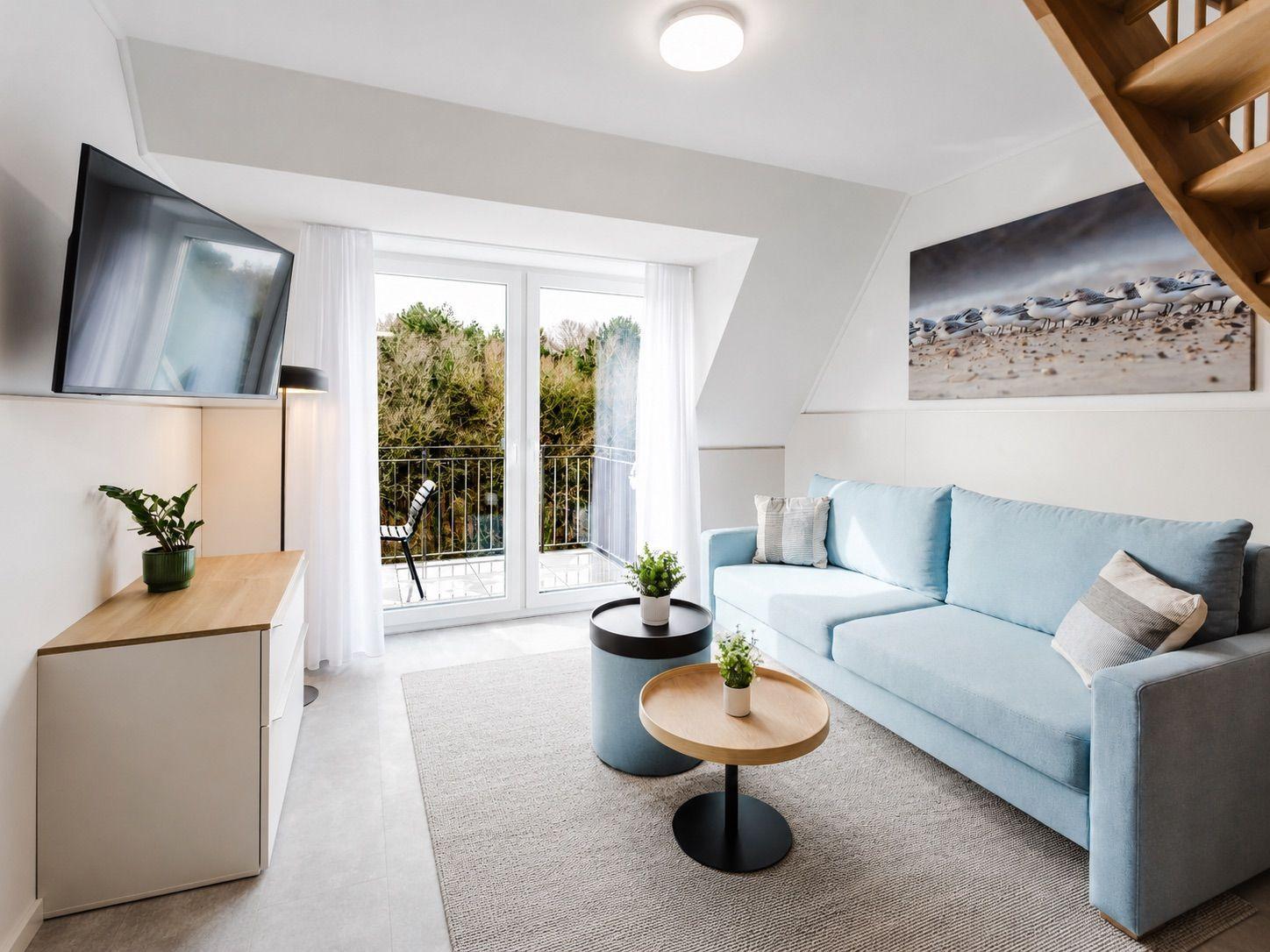 Wohnzimmer mit blauem Sofa, TV und Balkon. Holztreppe und Wandbild sichtbar.