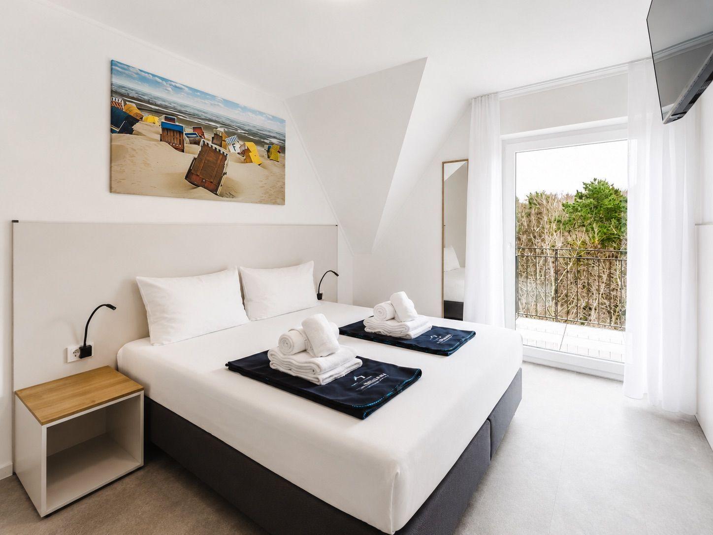 Schlafzimmer mit Doppelbett, Bademantel und Blick auf Balkon mit Bäumen.