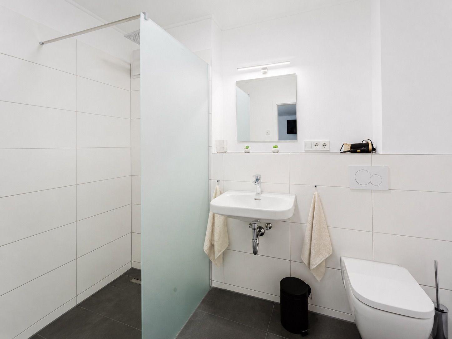 Badezimmer mit Dusche, Waschbecken und Toilette. Weiße Fliesen und dunkle Bodenfliesen.