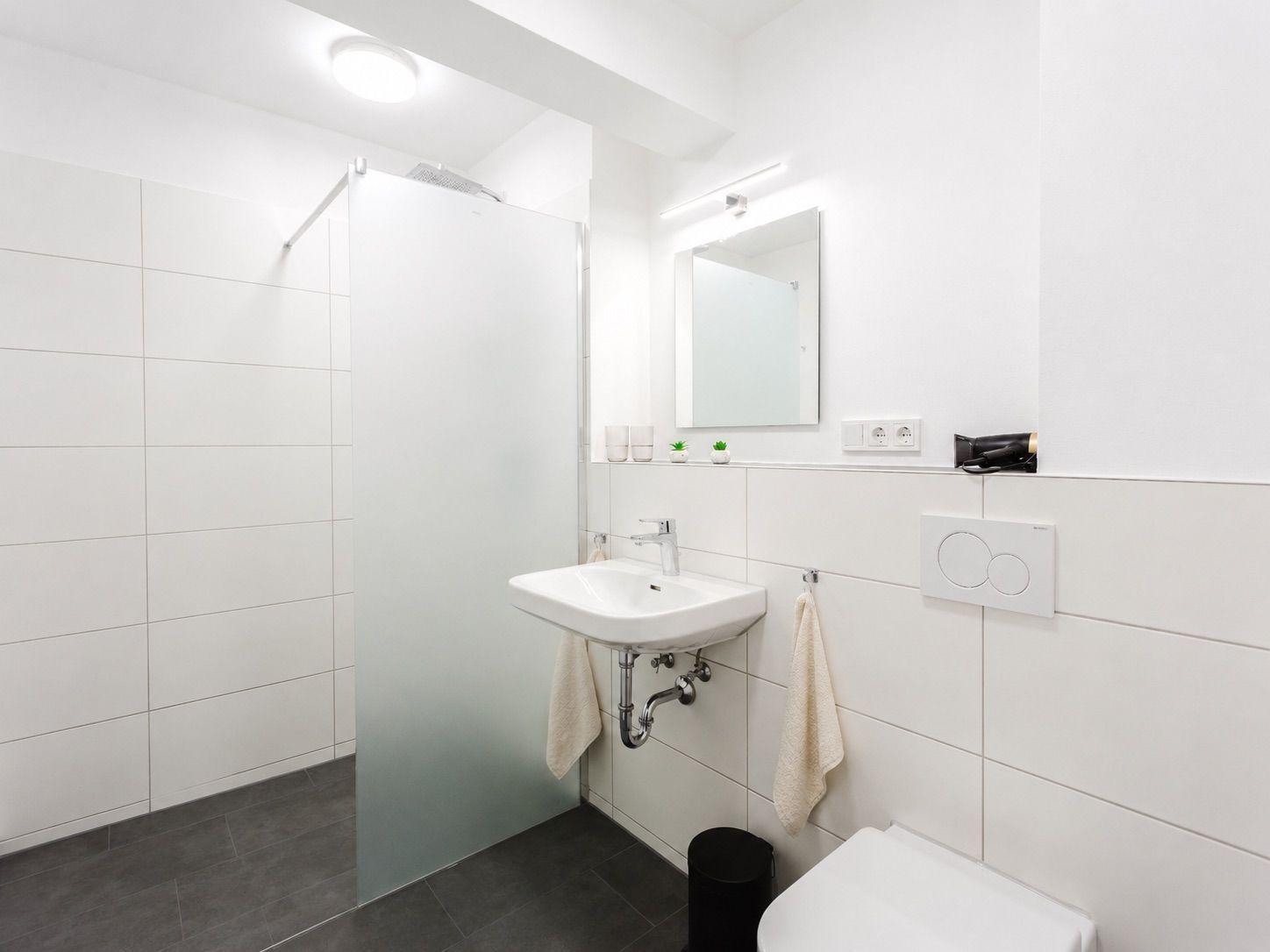Badezimmer mit Dusche, Waschbecken und Toilette. Weiße Fliesen und graue Bodenfliesen.