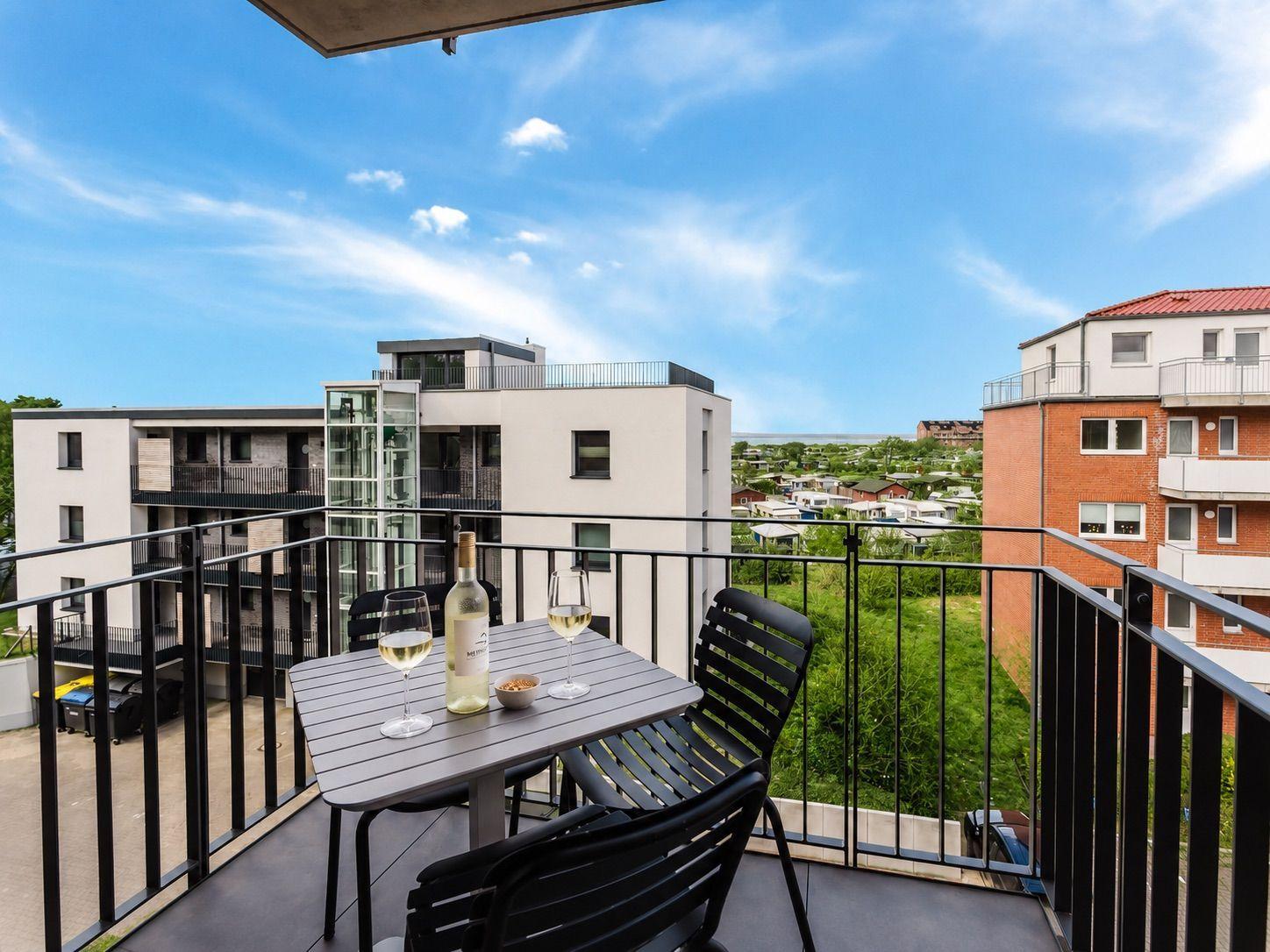 Terrasse mit Tisch und Stühlen. Hintergrund: Wohnhäuser und grüne Flächen unter blauem Himmel.