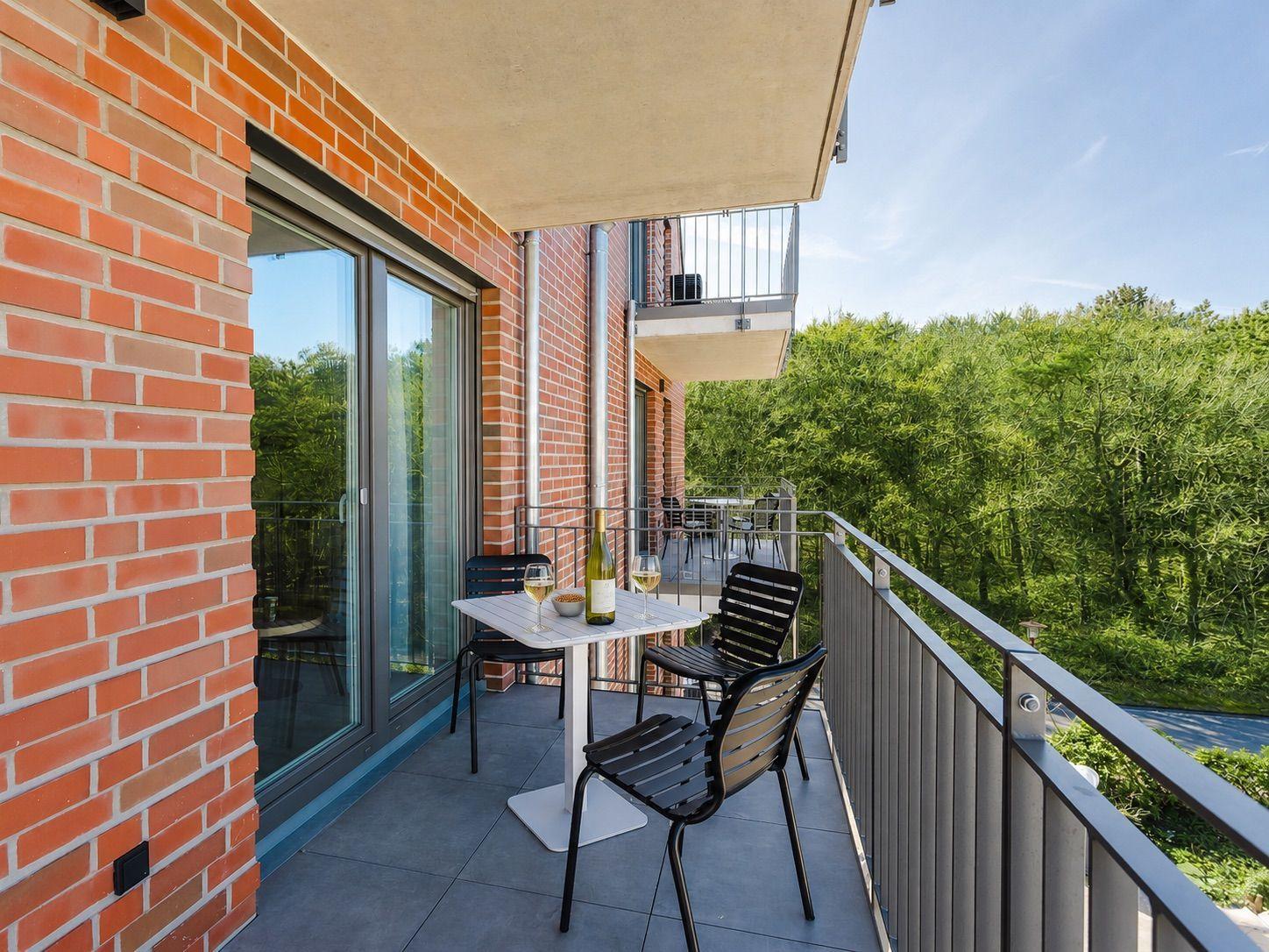 Terrasse mit Tisch, Stühlen und Weinglas. Blick auf grüne Bäume. Rotbraune Ziegelwand und Glasfront.
