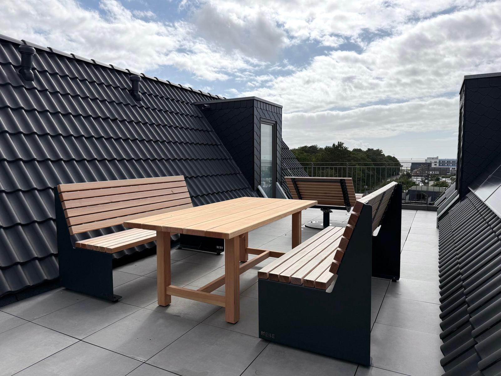 Terrasse mit Holztisch und Bänken, Blick auf Dächer und Landschaft.