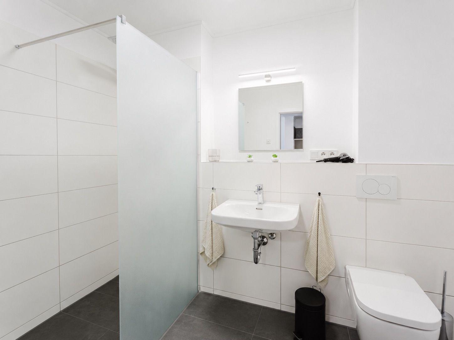 Badezimmer mit Dusche, Waschbecken und Toilette. Weiße Wände, grauer Boden.