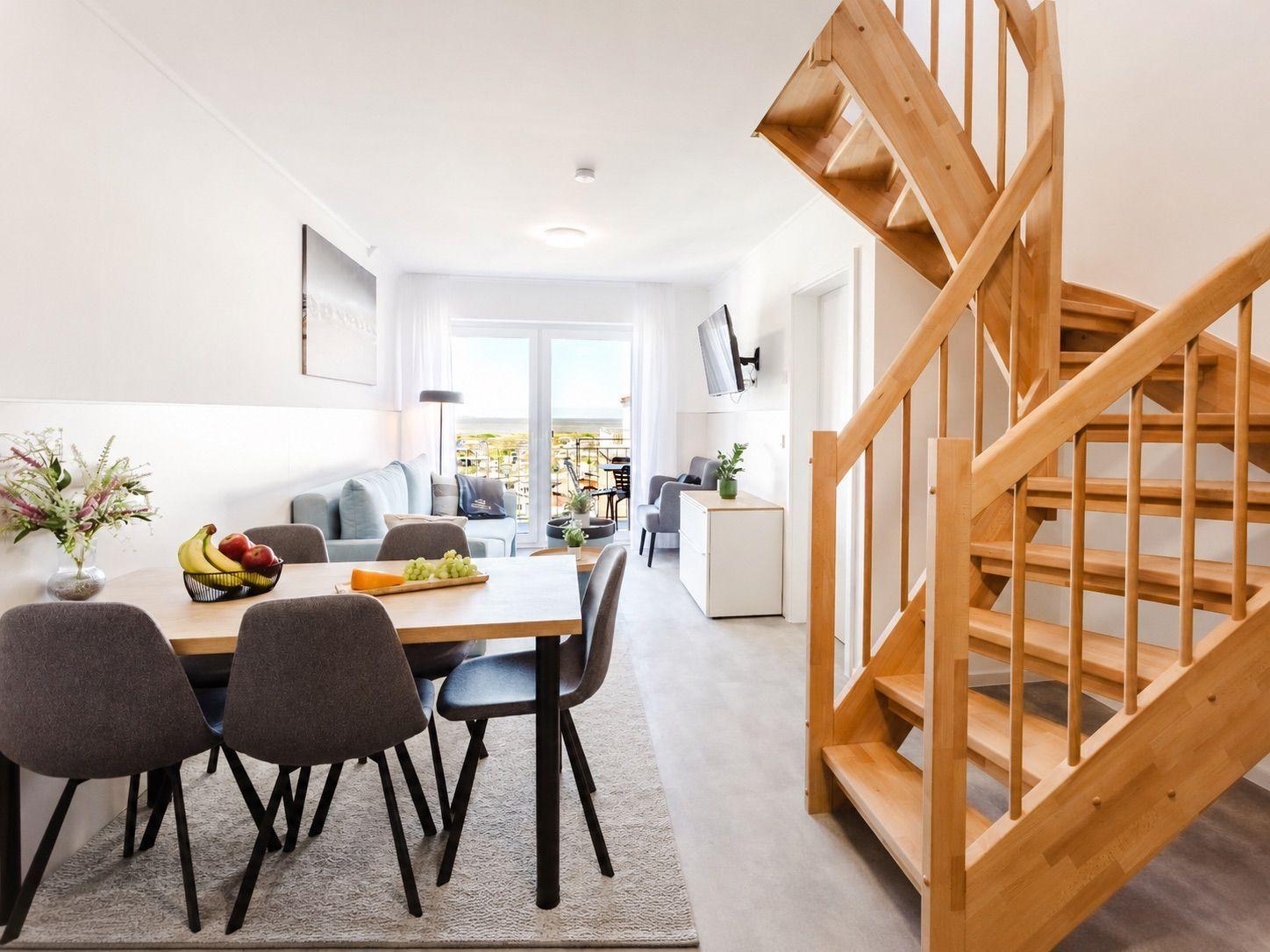 Essbereich mit Tisch und Stühlen, Wohnbereich mit Sofa und Balkon, Holztreppe zur oberen Etage.