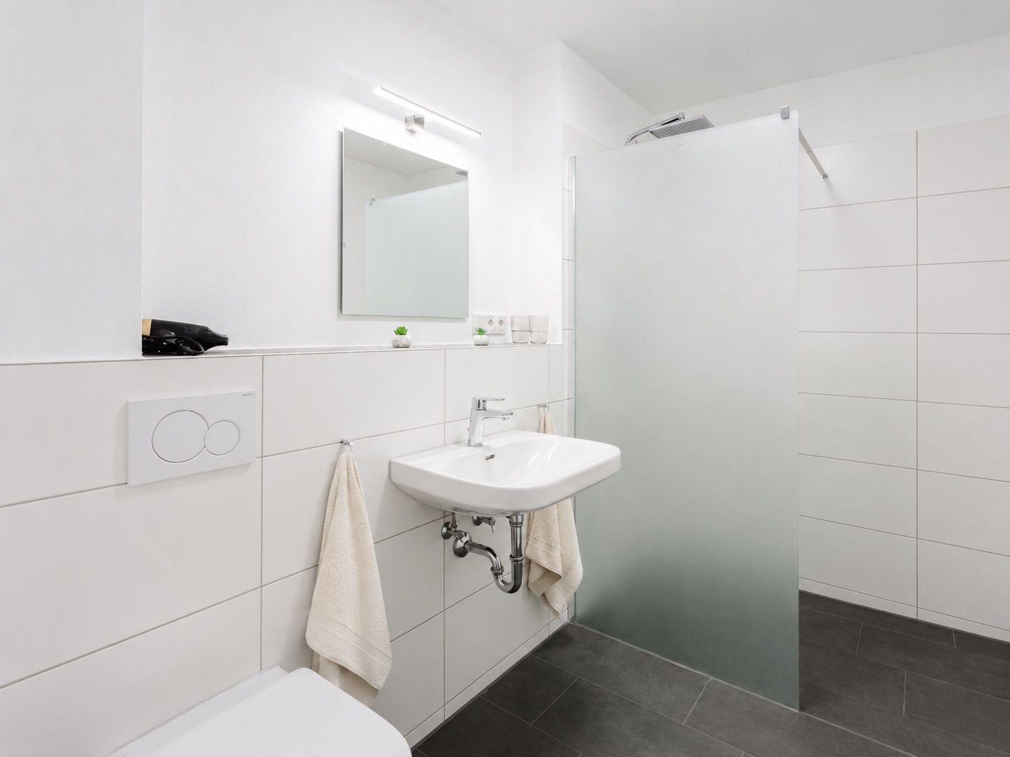 Badezimmer mit Dusche, Waschbecken und Spiegel. Weiße Fliesen und dunkler Boden.