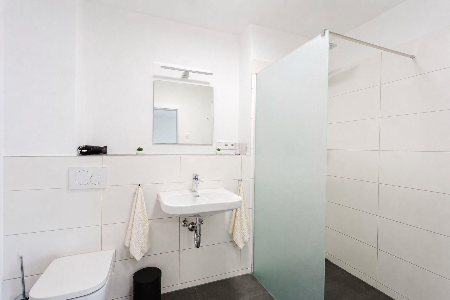 Badezimmer mit Dusche, Waschbecken und Toilette. Weiße Fliesen und Spiegel.