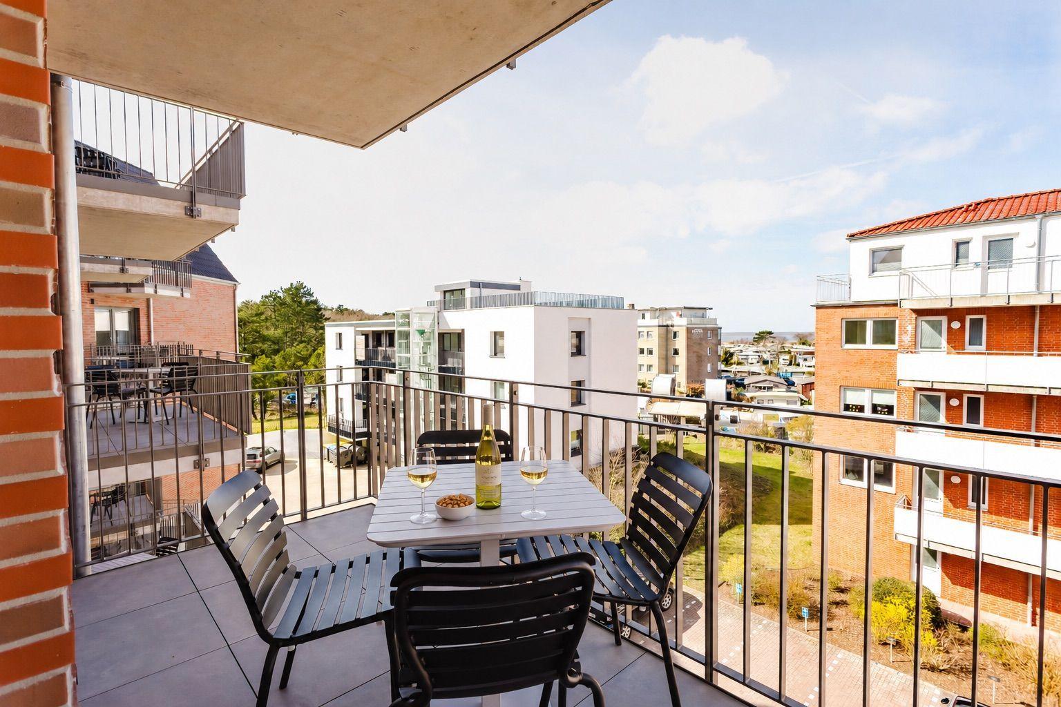Balcony with table, chairs, and wine bottle. View of modern buildings and green areas.