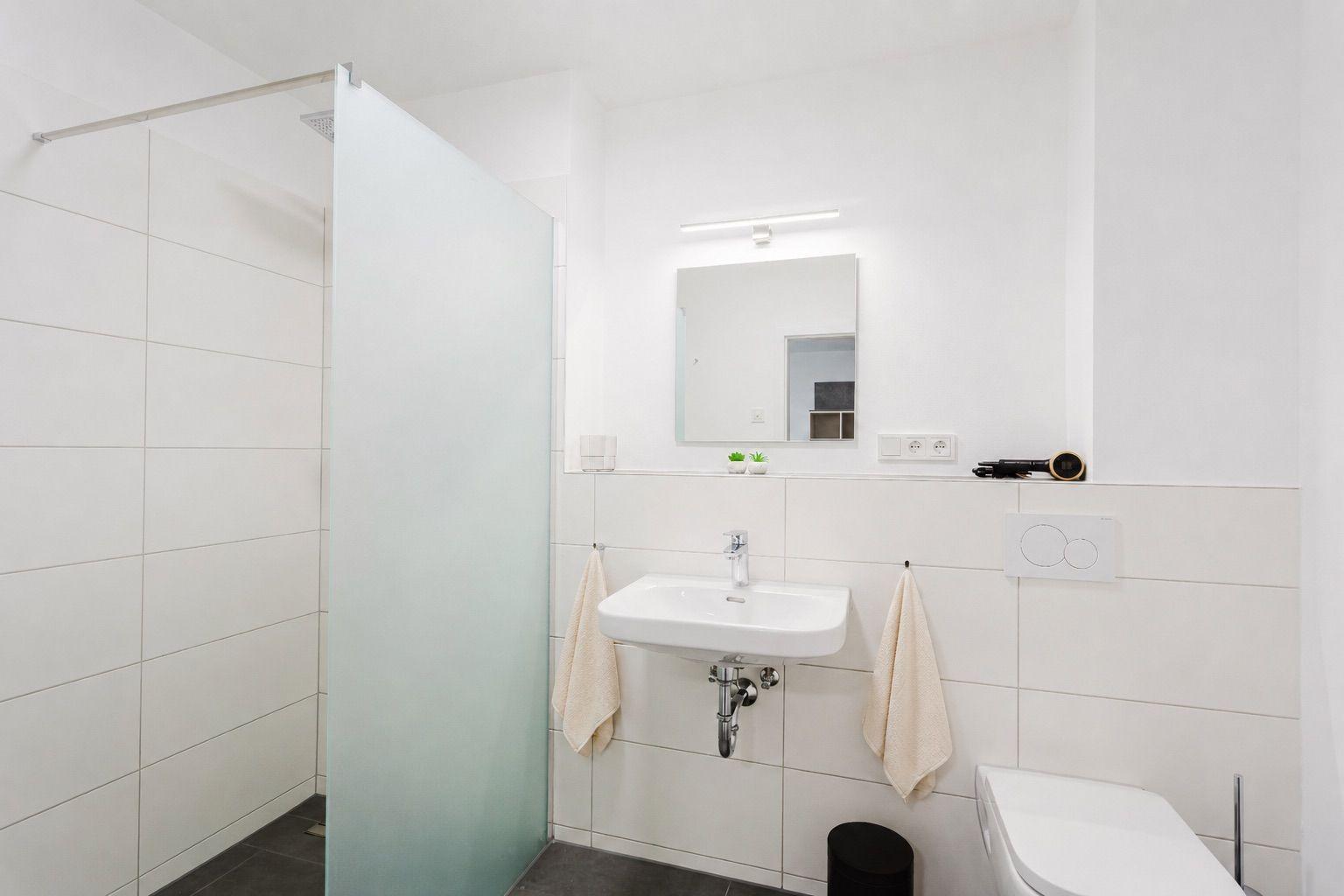 Badezimmer mit Dusche, Waschbecken und Toilette. Weiße Fliesen und Spiegel.