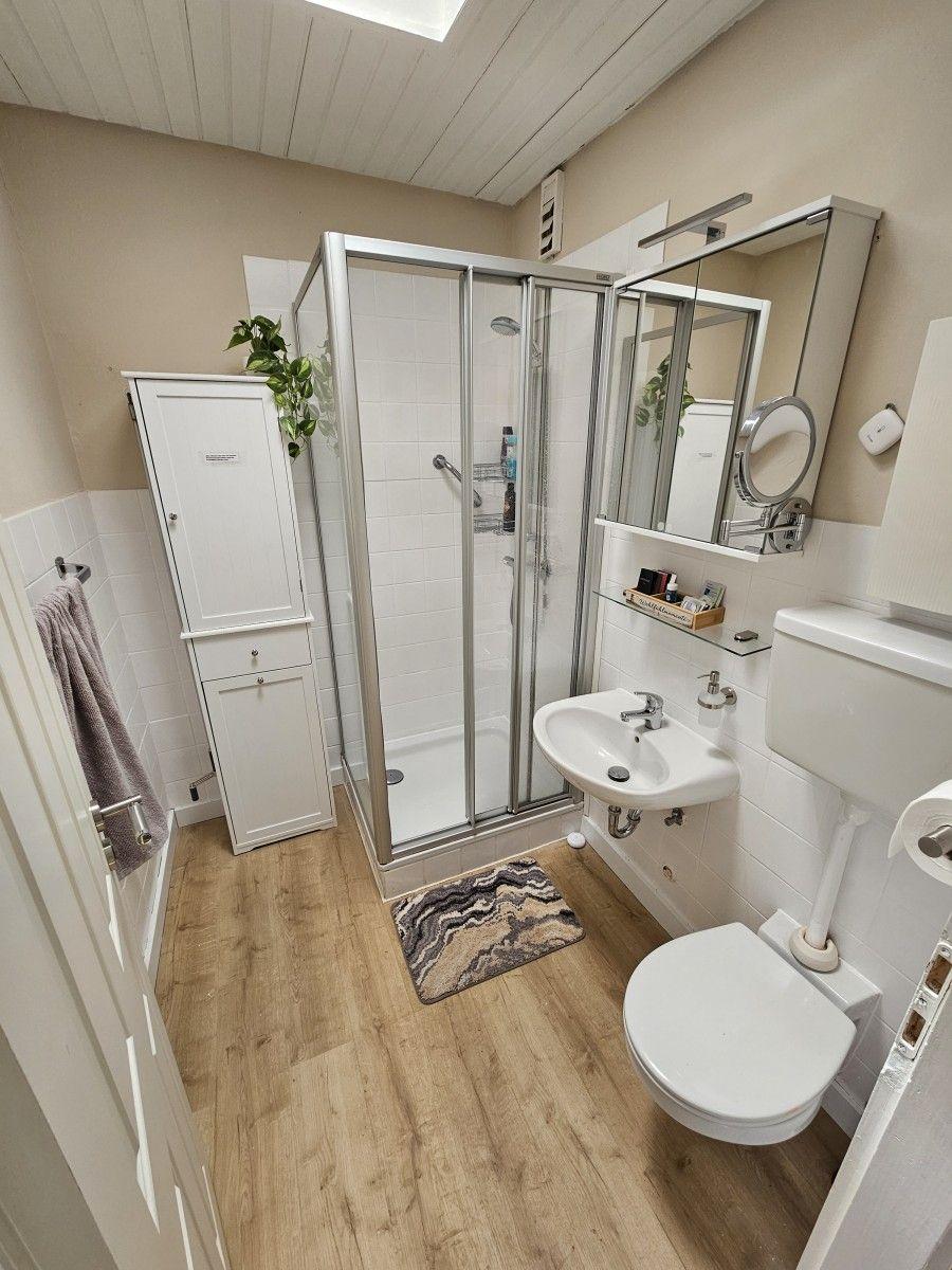 Bathroom with shower, sink, and toilet. Wooden floor and white cabinet.