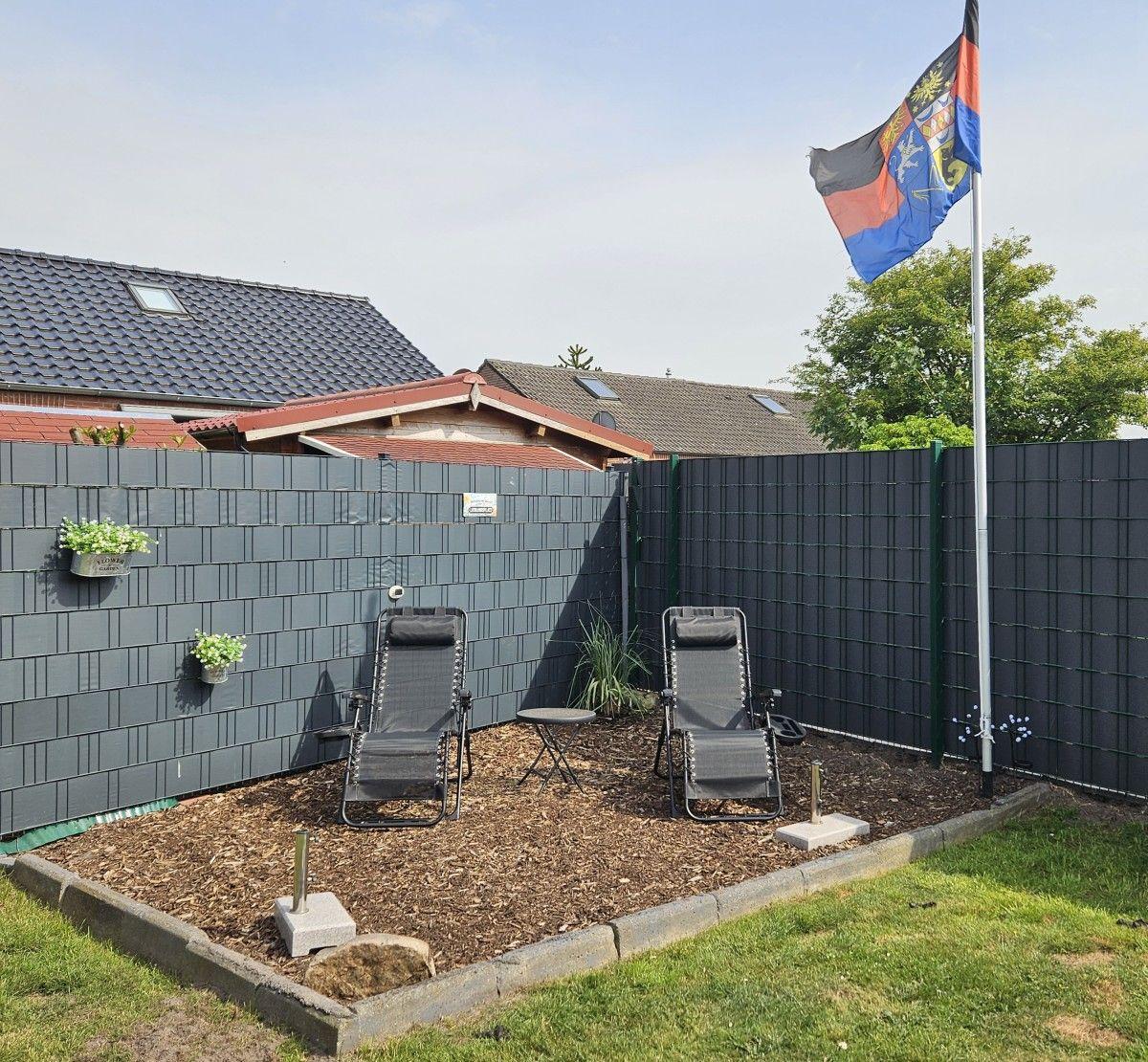 Backyard with lounge chairs, table, and flag. Fenced with dark wall and grass area.