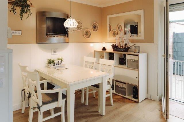 Dining room with white table, chairs, and kitchen area. A mirror and decor hang on the wall.