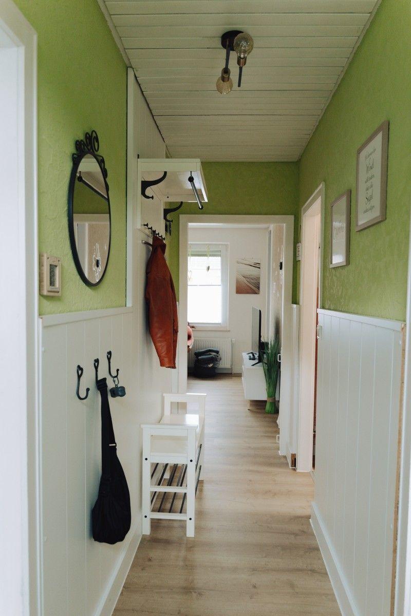 Hallway with green walls, white wainscoting, and wooden floor. Hooks and a mirror are on the wall.