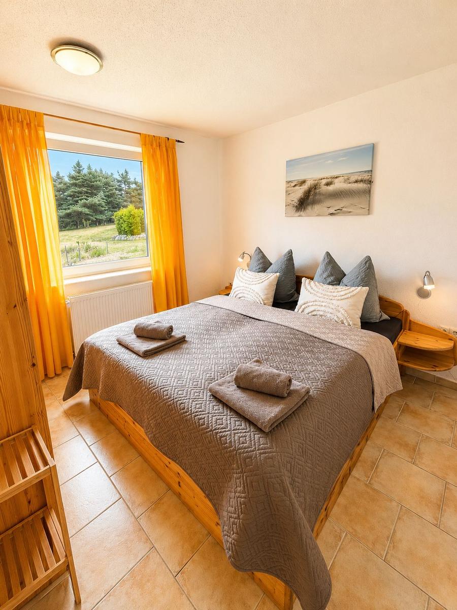 Schlafzimmer mit Bett, Fenster und Wandbild. Holzmöbel und gefaltete Handtücher.