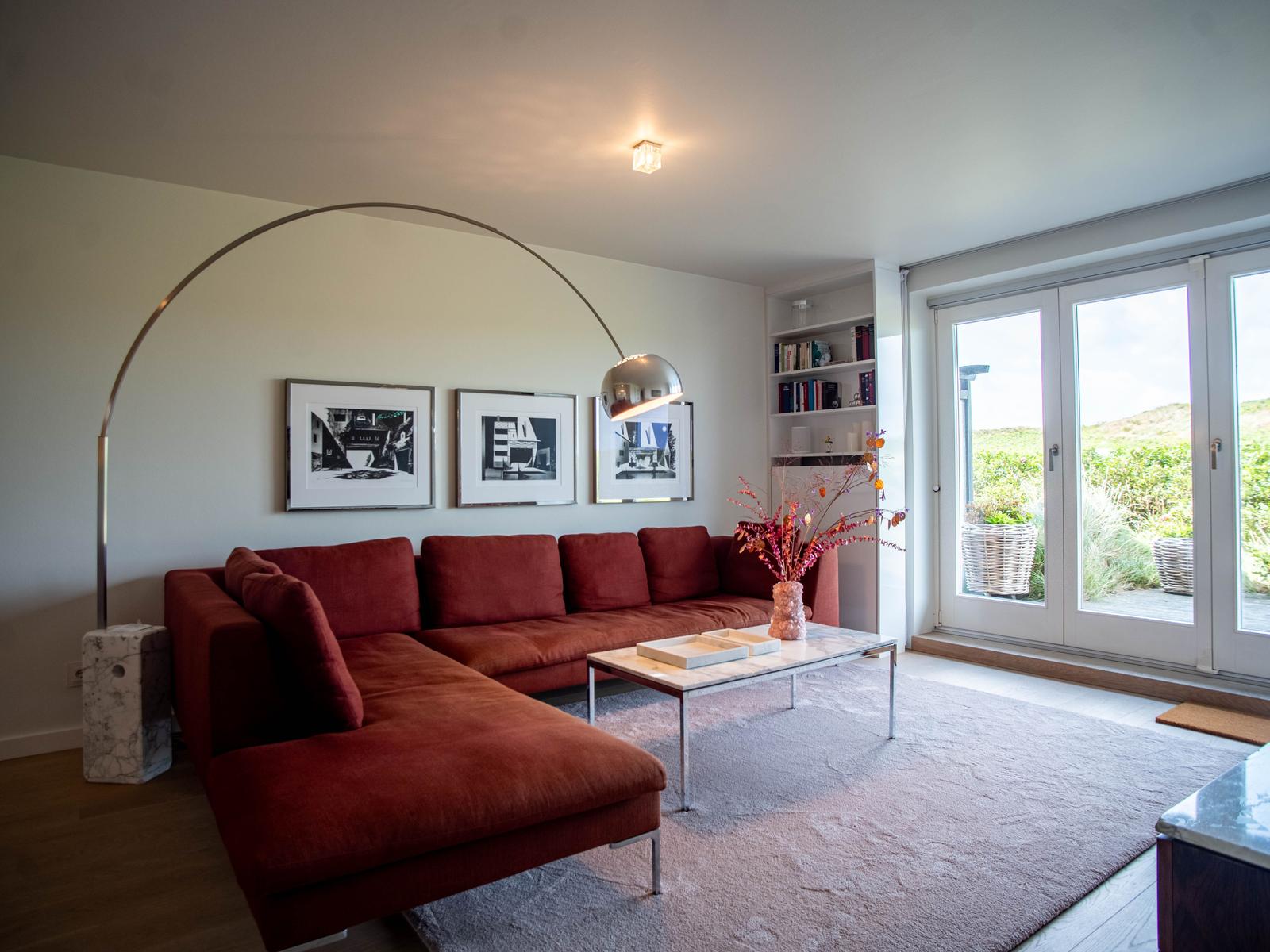 Ein Wohnzimmer mit rotem Sofa, Glas-Tisch und Bücherregal. Durchgang zur Terrasse mit Blick auf Grünflächen.