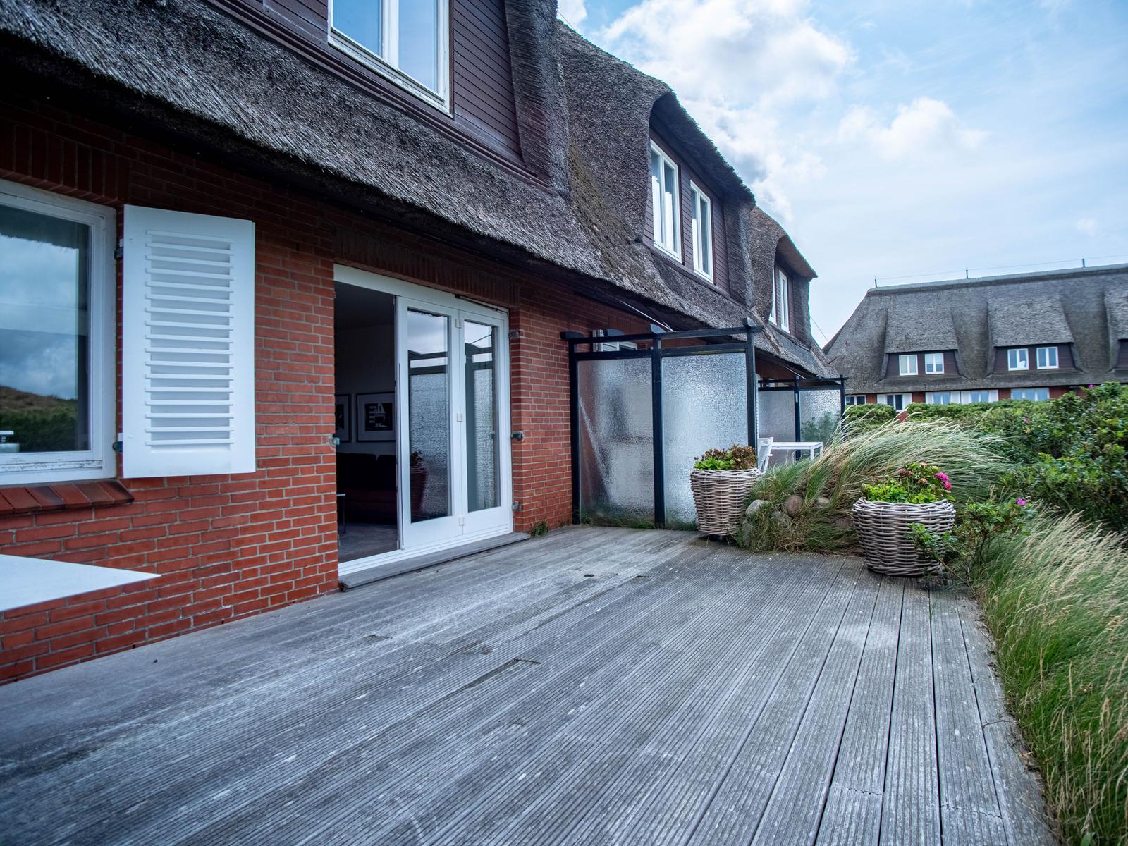 Holzterrasse mit Blick auf ein Haus mit strohgedecktem Dach und Pflanzen in Körben.