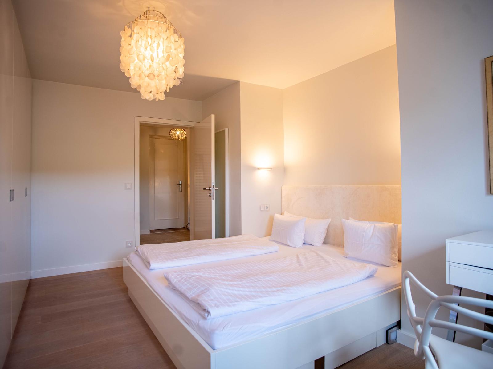 Schlafzimmer mit weißem Bett, Holzboden und Deckenleuchte. Tür führt in einen Flur.