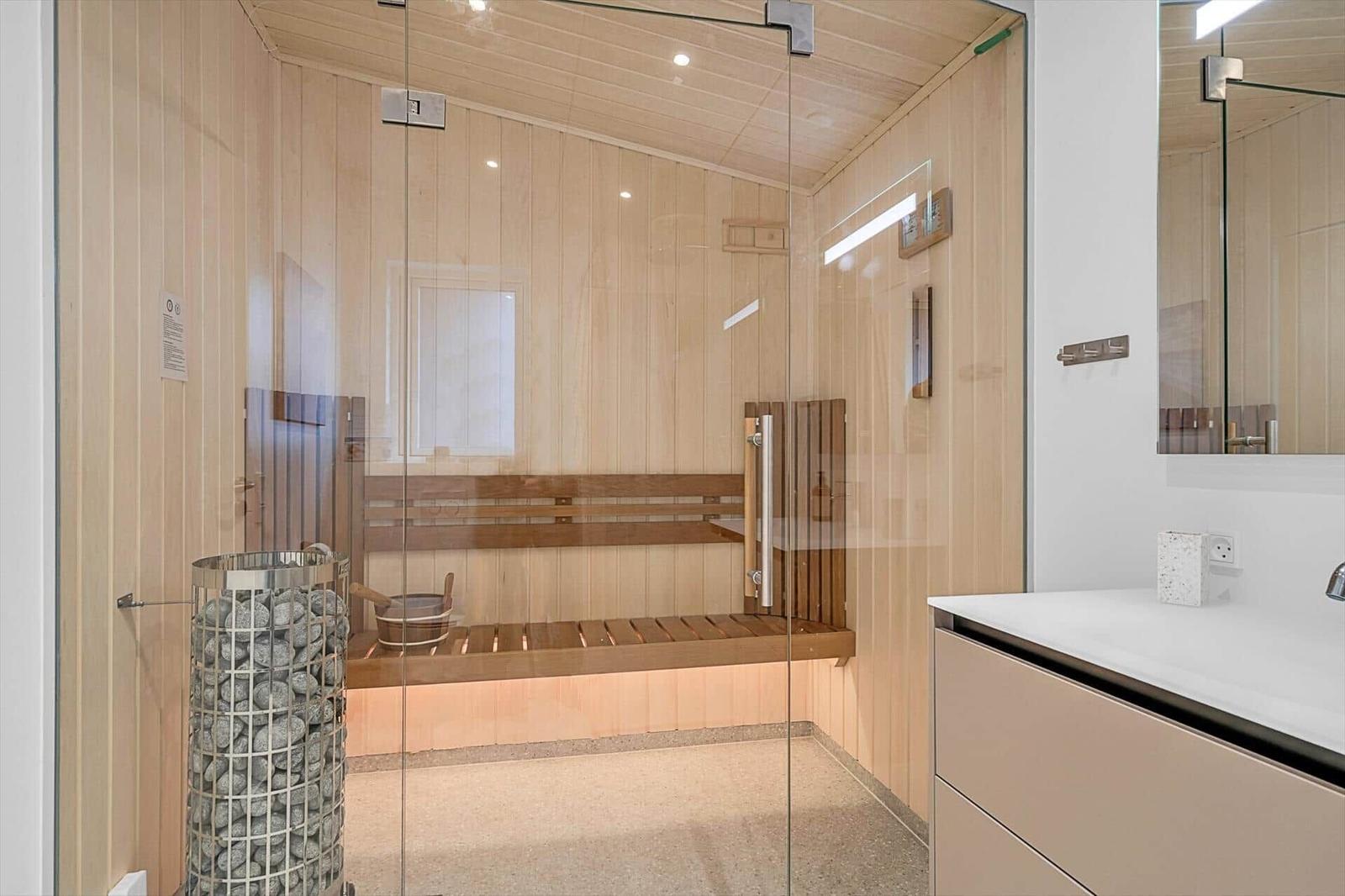 Sauna mit Holzwand, Sitzbank und Steinbehälter. Glaswand trennt Raum von Bad.