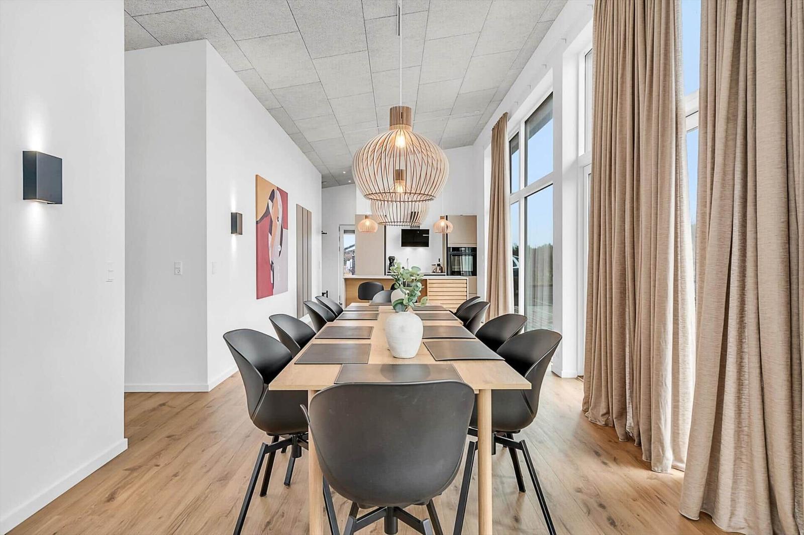 Esszimmer mit langem Tisch, schwarzen Stühlen und hängenden Lampen. Holzboden und große Fenster.