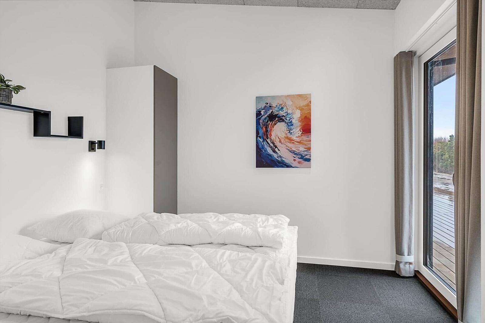 Schlafzimmer mit weißem Bett, Wandbild und Fenster zur Terrasse.