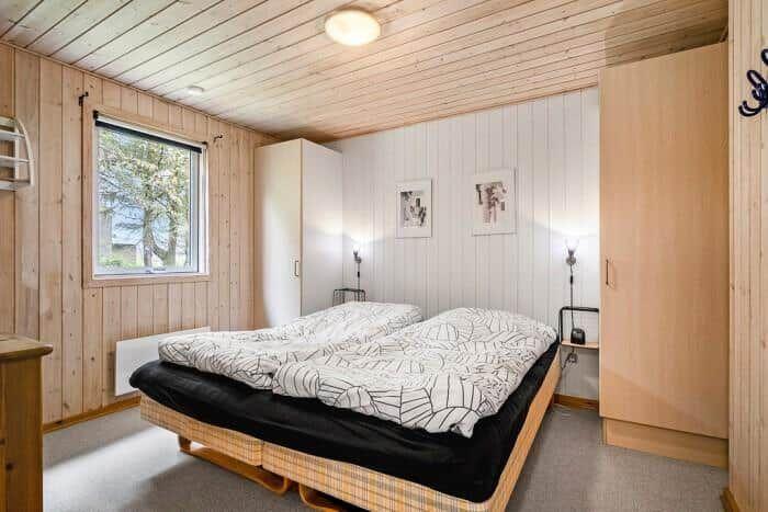 Schlafzimmer mit Doppelbett, Holzwand und Fenster. Hängende Bilder an der Wand.