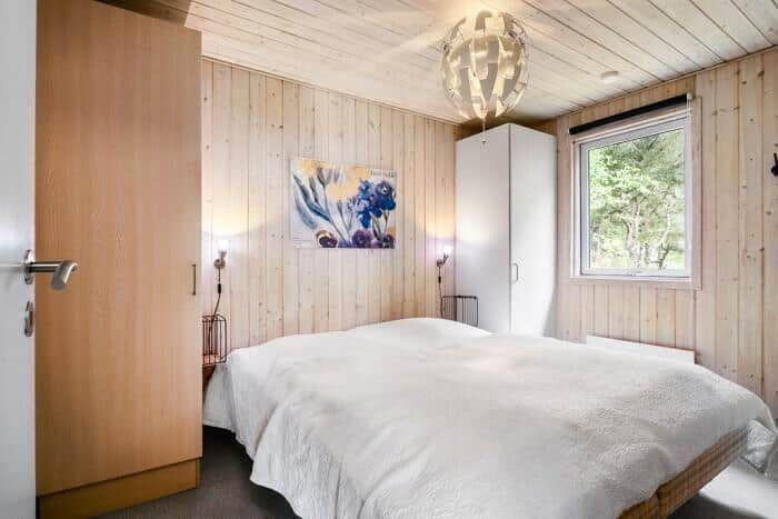 Schlafzimmer mit Holzwand, Bett, Kleiderschrank und Fenster mit Blick auf Bäume.