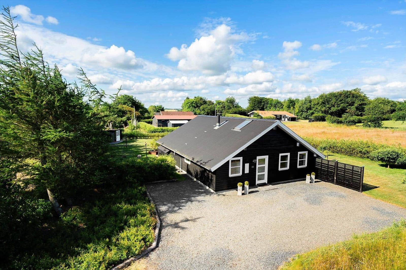 Schwarzes Haus mit grauem Dach, umgeben von Grünflächen und Bäumen unter blauem Himmel.