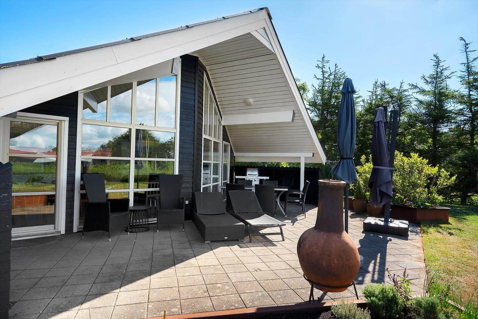 Terrasse mit Sitzgelegenheiten und Grillplatz. Hintergrund: Wald und Haus mit Glasfassade.